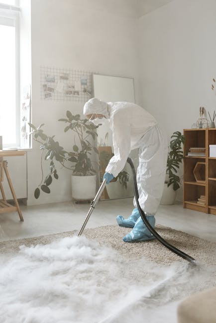 A person wearing orange work overalls vacuuming a modern living room with a yellow industrial carpet cleaning machine. The room features a grey fabric sofa with beige pillows, a black metal coffee tab